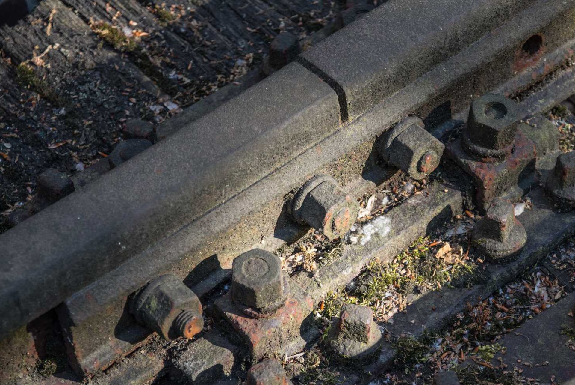 Fot. a. Zdjęcie przedstawia złączenie dwóch żelaznych szyn kolejowych. Dwa proste odcinki podłużnych i grubych elementów stanowiących szyny kolejowe przyłożone są do siebie w taki sposób, że złącze przedłuża je. Szyny położone są na grubych, drewnianych belkach częściowo wkopanych w ziemię, przy  czym belki położone są poprzecznie w stosunku do długości szyn. Szyny połączone są żelaznymi mocowaniami w postaci prostopadłościennych płyt żelaznych, które skręcono z szynami przy pomocy żelaznych sześciokątnych śrub. Pomiędzy przedłużającymi się szynami widoczna jest wyraźna przerwa. Obecność tych przerw w torowisku odczuwalna jest podczas jazdy w postaci charakterystycznych stuków. Wielkość przerwy pomiędzy przedłużającymi się szynami zmienia się w zależności od temperatury powietrza ze względu na rozszerzalność cieplną. W wyższych temperaturach szyny zwiększają swoją objętość, a wielkość przerwy między szynami zmniejsza się.