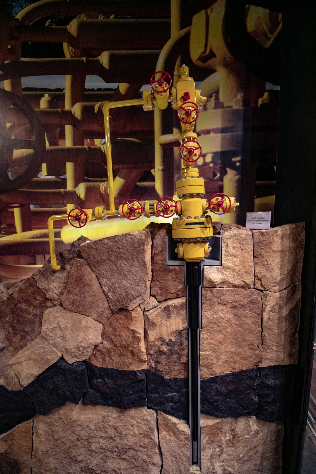Na pionowym zdjęciu widoczny jest przekrój przez odwiert z zamontowaną głowicą eksploatacyjną. Na pierwszym planie widoczny jest przekrój przez skałę widoczny jako trzy warstwy popękanych skał – od góry brązowego koloru, czarnego koloru i brązowego koloru. W prawej części widoczny odwiert jako wydłużony element zwężający się w 4 stopniach, przy czym w obwiedni tego elementu widoczne szare ścianki, zaś wypełnienie jest czarnym wrzecionowatym elementem wydłużonym w pionie. W górnym rozszerzeniu zlokalizowana głowica eksploatacyjna o korpusie koloru żółtego składająca się z pionowego elementu złożonego z dwóch połączonych ze sobą płaskich elementów na których obwiedniach widoczne są główki śrub, powyżej ze zwężenia od którego odchodzą w stronę lewą pojedyncza zasówa z widocznym czerwonym kołem, zaś z lewej strony dwie zasuwy umiejscowione poziomo w lewą stronę, następnie zwężenie od którego odchodzi w górę cienka pionowa rura koloru żółtego, następnie zasuwa z kołem oraz kolano ze zwężonej rury prowadzące pionowo w dół do pierwszej warstwy skał. Powyżej rozwidlenia w głównym pionowym elemencie głowicy znajduje się podwójny połączony śrubowo element oraz zespół dwóch zasów z czerwonymi kołami do ich obsługi, umiejscowionych jedna nad drugą pionowo, prowadzące do prostokątnego wydłużonego w pionie elementu zawierającego dwa małe elementy od góry i od prawej strony. Od lewej strony tego elementu widoczna jest zasuwa z czerwonym kołem, połączenie śrubowe dwóch kołnierzy oraz rura pozioma, przechodząca w kolano skierowane w dół, prowadząca do łączenia z dolną lewą odnogą głowicy. W tle widoczne zdjęcie orurowania w kolorze żółtym oraz karteczka z podpisem modelu z nieczytelnym tekstem. 