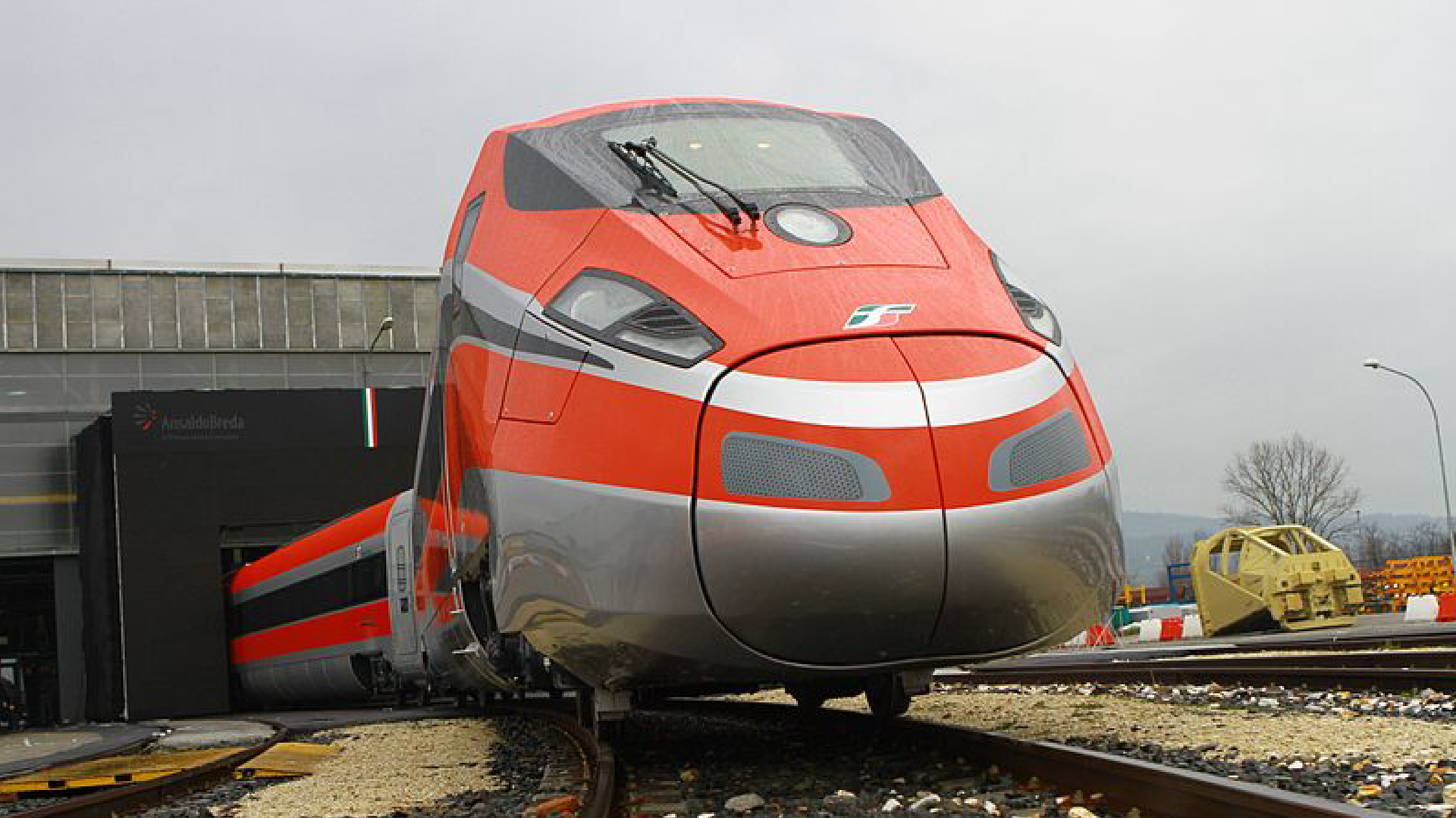 Zdjęcie przedstawia zbliżenie na przód nowoczesnego włoskiego pociągu. Pociąg znajduje się na torach, wyjeżdża z hangaru.