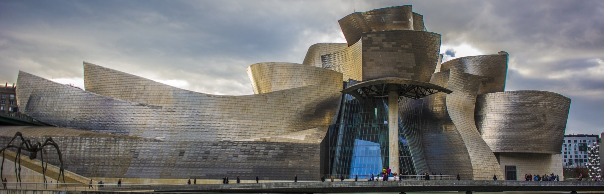 Ilustracja o kształcie poziomego prostokąta przedstawia fotografię Muzeum Guggenheima Franka O. Gehry’ego. Ukazuje budynek o rozczłonkowanej bryle i masywnym kształcie, położony nad akwenem oraz na tle architektury miejskiej. Nad miastem rozciąga się zachmurzone niebo.