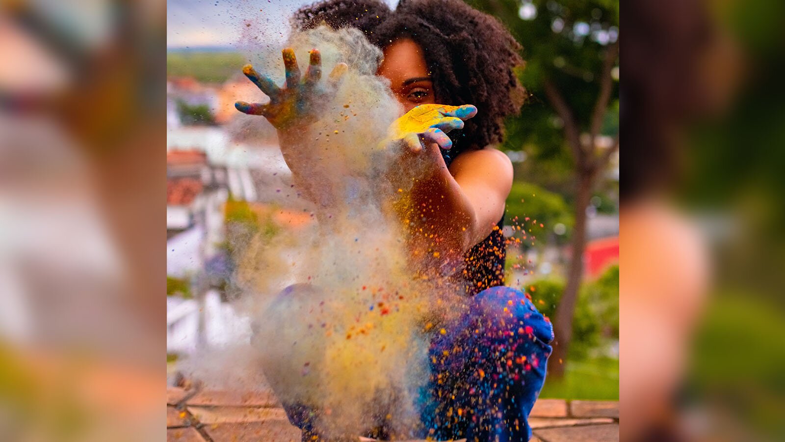 Zdjęcie ukazuje młodą kucającą kobietę, która rzuca kolorowym proszkiem w stronę aparatu. Jej dłonie są umazane.