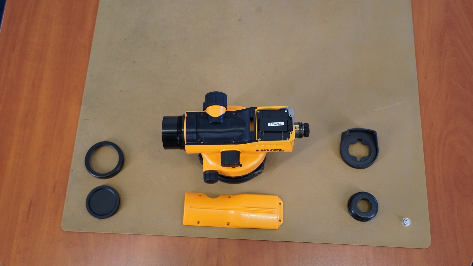 Zdjęcie przedstawia urządzenie widziane od góry. Zdemontowano z niego górną osłonę oraz obudowę okularu. Obok niwelatora umieszczono zdemontowane elementy: obudowę okularu złożoną z dwóch części oraz górną osłonę, obręcz obiektywu i jego przysłonę.