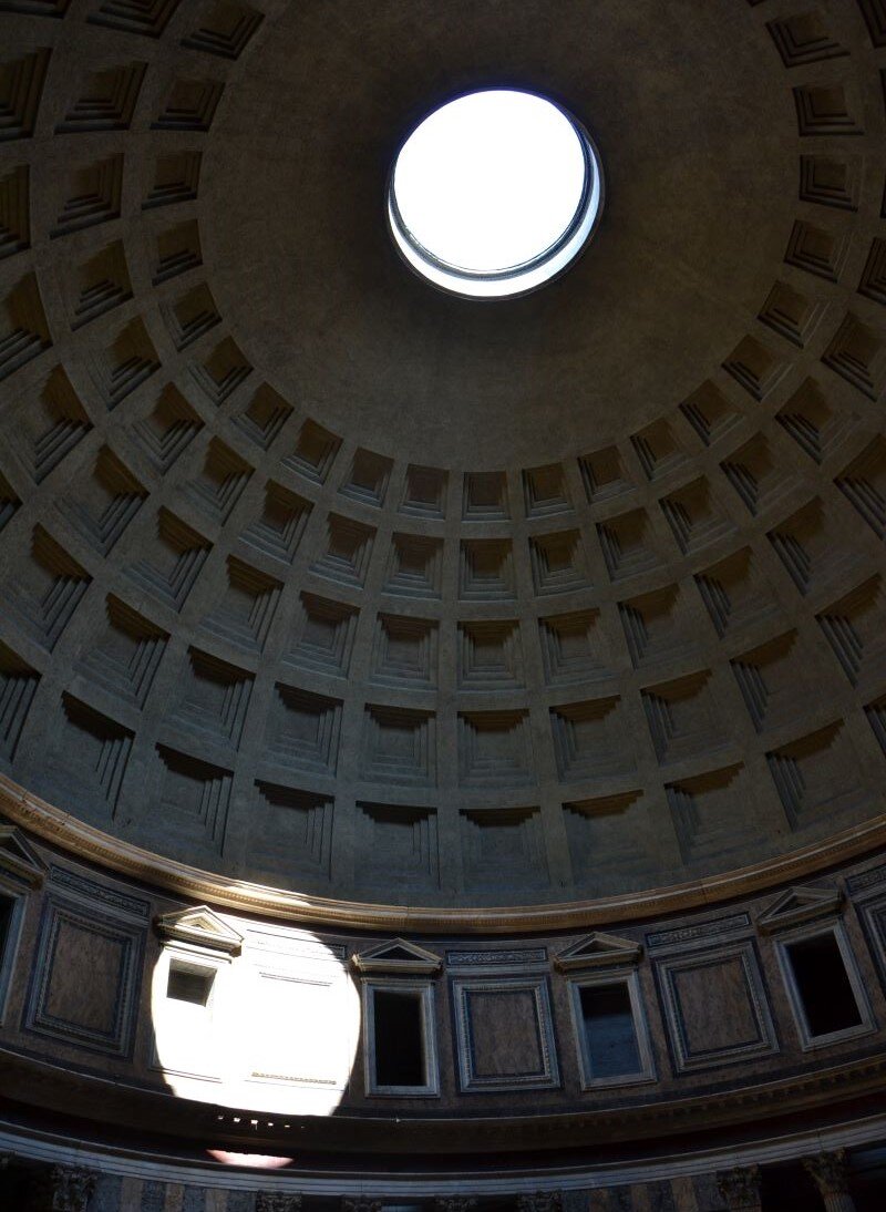 Ilustracja przedstawia fotografię fragmentu wnętrza. A kadrze znajduje się widok kopuły z kasetonami i jednym okrągłym otworem, przez który wpada białe światło. Całość fotografii jest ciemna, tylko światło w kopule oraz światło o takim samym kolorze i kształcie okręgu, padające na fragment ściany powoduje, że wnętrze nie jest pogrążone w ciemności. Widoczny jest silny kontrast między światłem i cieniem.