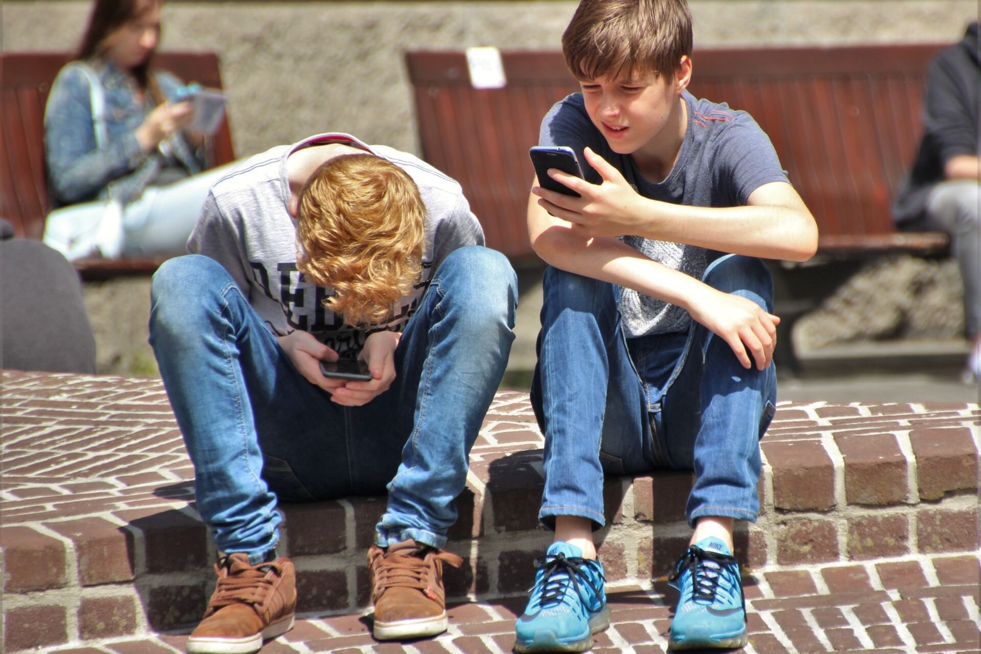 Zdjęcie przedstawia dwóch chłopców siedzących na chodniku z telefonami w rękach. Jeden z nich jest głęboko skupiony nad telefonem trzymanym między kolanami, podczas gdy drugi jest bardziej wyprostowany, ale także skoncentrowany na swoim telefonie. W tle widoczne są inne osoby siedzące na ławkach, również korzystające z telefonów.