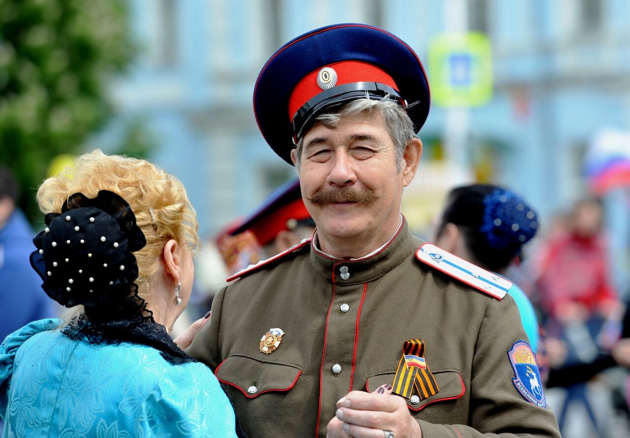 Fotografia kolorowa przedstawia kobietę tańczącą taniec wolny ze starszym mężczyzną, który ubrany jest w strój przypominając stalina. Charakterystyczna czapka z krótkim daszkiem jest przekrzywiona na jego głowie, spod niej wystają siwe włosy. Mężczyzna uśmiecha się lekko, mrużąc mocno oczy. Uniform wojskowy, który nosi nie wyróżnia się niczym, poza podobieństwem do ubioru stalina, posiada na ramieniu naszywkę z jeleniem. Widoczna jest jedynie głowa i ramiona kobiety, od tyłu. Widać włosy związane z czarną ozdobą z wieloma kuleczkami z tyłu głowy. Tło rozmazane.