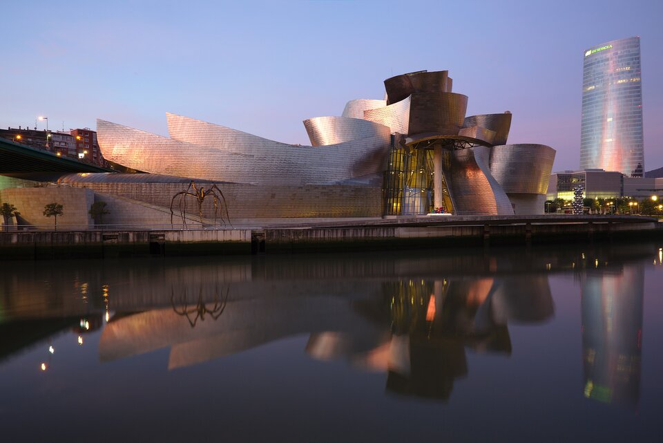 Zdjęcie ukazuje budynek Muzeum Guggenheima w Bilbao w Hiszpanii, sfotografowany o zmierzchu na tle fioletowego nieba. Budynek ma z zakrzywione, błyszczące powierzchnie, które wyglądają jak wykonane z metalu. Jego kształty odbijają światło. Muzeum znajduje się nad brzegiem rzeki,  na powierzchni której odbijają się budynek i niebo. W tle, za muzeum, rozciąga się panorama miasta. 