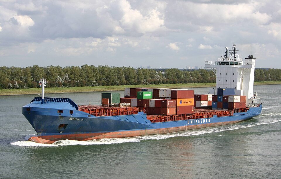 Zdjęcie przedstawia kontenerowiec na rzece. Jest on niebieski. Na pokładzie znajdują się kontenery. Z tyłu posiada wieżę z miejscem sterowania. W tle na brzegu rosną drzewa.