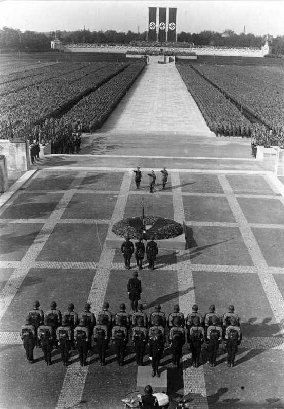 Zdjęcie przedstawia zjazd partii NSDAP w Norymberdze w 1935 r. Na pierwszym planie widać żołnierzy stojących w dwuszeregu na ogromnym placu, tyłem do obiektywu. Przed nimi stoi jeden żołnierz , a przed nim jeszcze trzej żołnierze w szeregu, którzy trzymają w dłoni flagę. Przed nimi znajduje się okrągły klomb. Przodem do nich i do obiektywu stoją trzej ludzie z wyciągniętymi do przodu wyprostowanymi prawymi rękami w geście Heil Hitler. W tle widoczna jest długa aleja z białym murem na końcu, nad którym znajdują się trzy duże flagi ze swastyką na białym tle. Po obu stronach alei widoczne są rzesze ludzi stojące w równych rzędach.