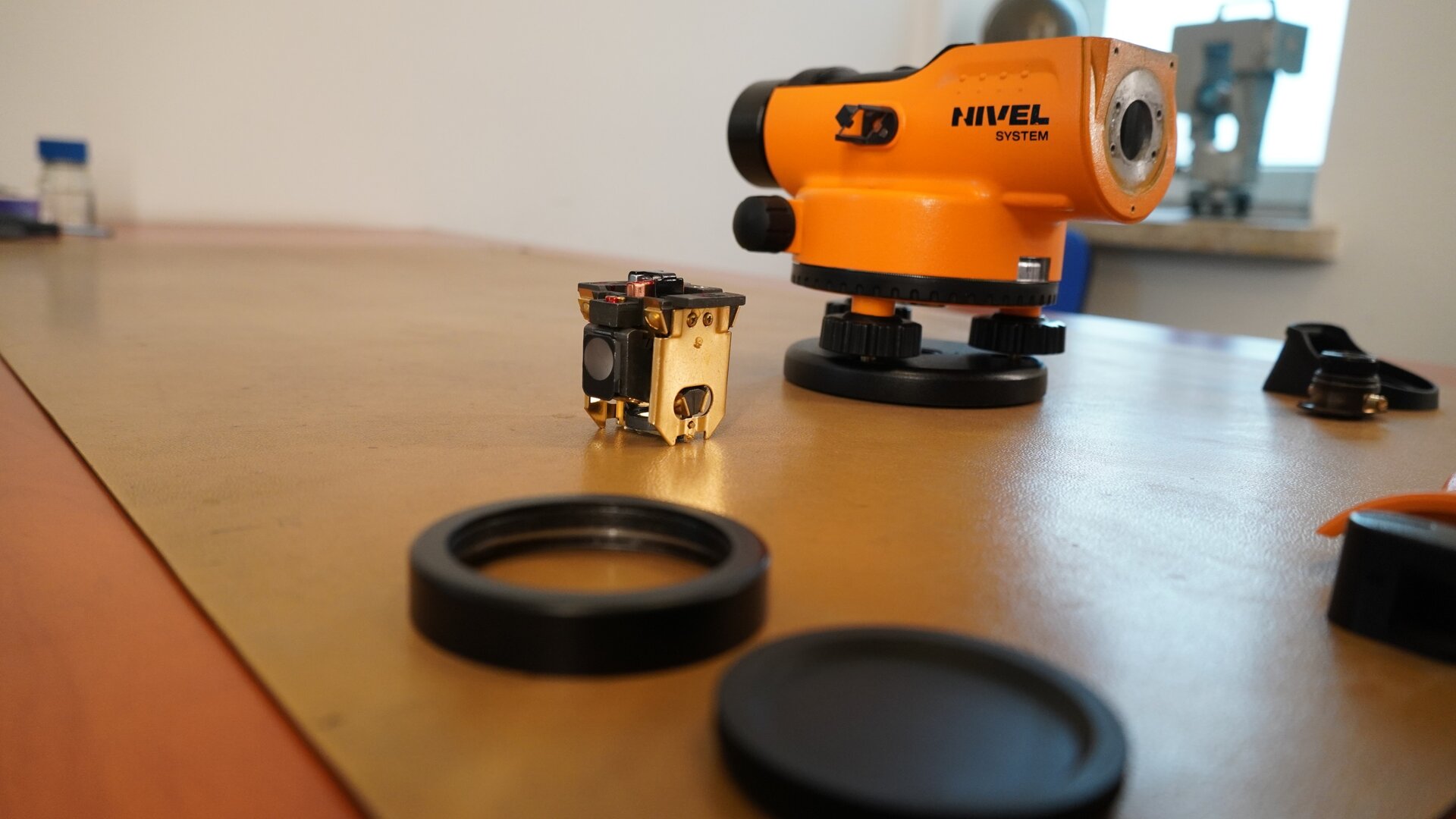 Na zdjęciu przedstawiono niwelator po demontażu okularu, jego obudowy oraz kompensatora. Zdemontowane części ustawiono przed instrumentem pomiarowym.