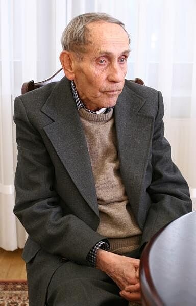 Na zdjęciu widnieje siedzący przy stole starszy mężczyzna, ma krótkie włosy, głęboko osadzone oczy oraz wyraźne kości policzkowe. Ubrany jest w marynarkę, pod którą ma sweter oraz wystający kołnierzyk od koszuli w kratę.