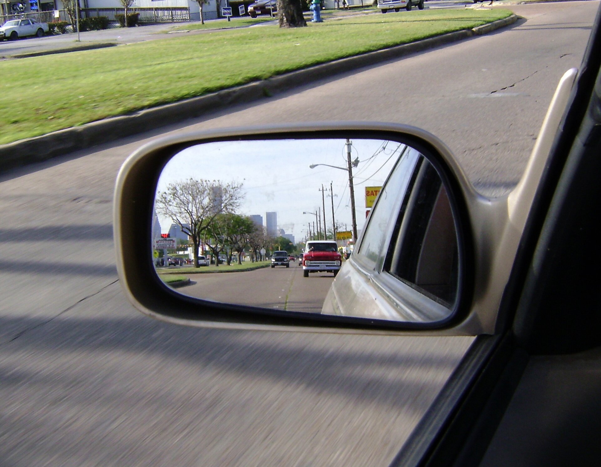 Zdjęcie przedstawia lewe lusterko samochodowe. W odbiciu widać inne auta znajdujące się na drodze z tyłu. Widoczny fragment samochodu, szary asfalt ulicy i pas zieleni z fragmentem pnia rosnącego drzewa.