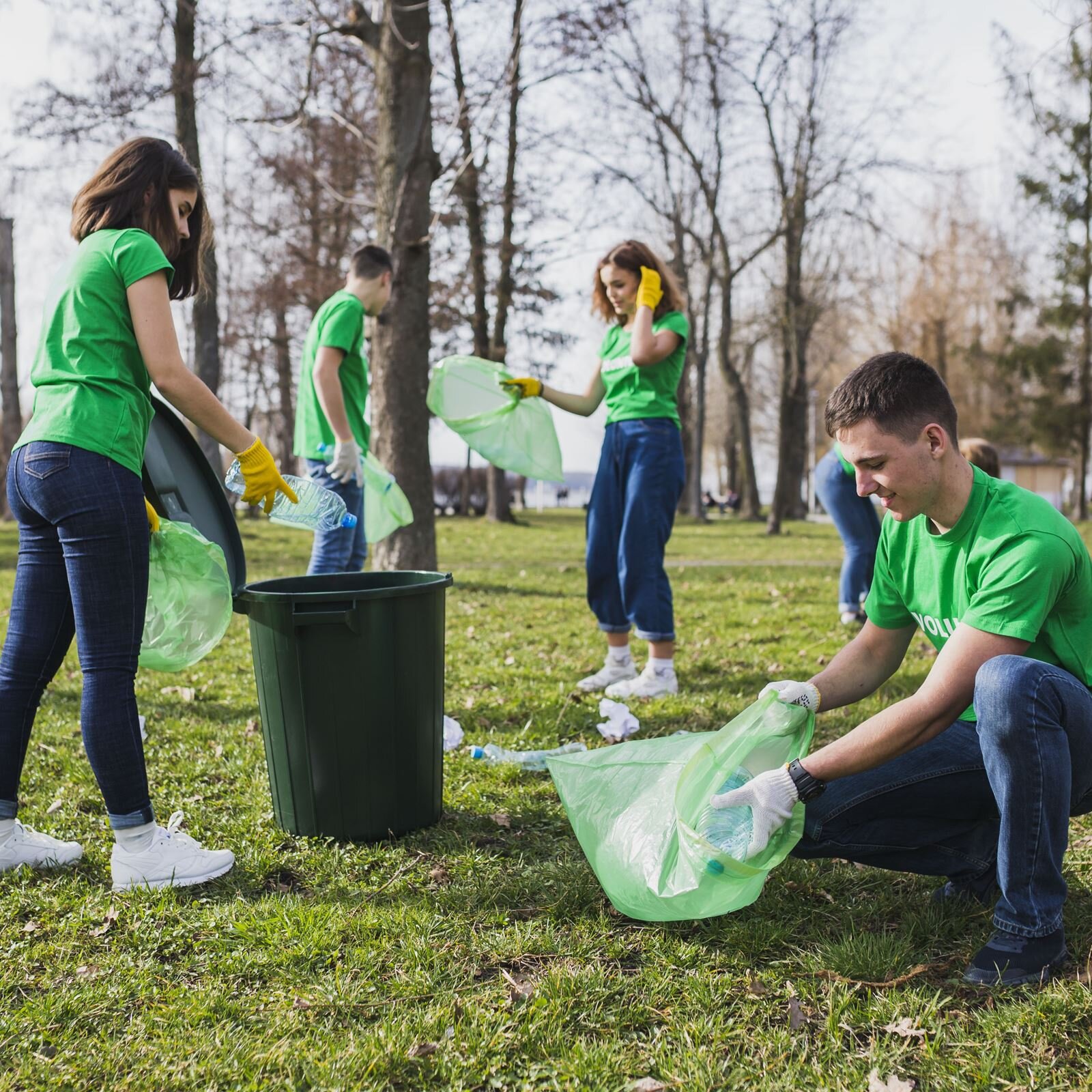 Zdjęcie przedstawia grupę młodych aktywistów, ubranych w zielone koszulki, niebieskie spodnie i rękawiczki ochronne, zbierających śmieci w parku. Aktywiści znajdują się w parku i zbierają do worków porozrzucane śmieci.
