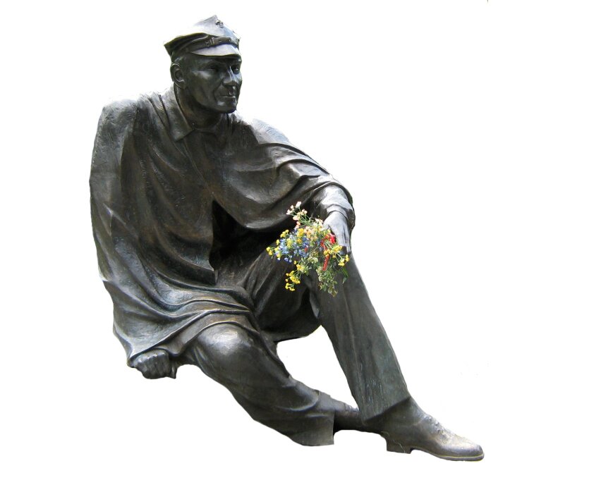 Kliknij, aby powiększyć. Zdjęcie pomnika, przedstawia siedzącego mężczyznę w czapce i pelerynie harcerskiej. W dłoni ma bukiecik polnych kwiatów.