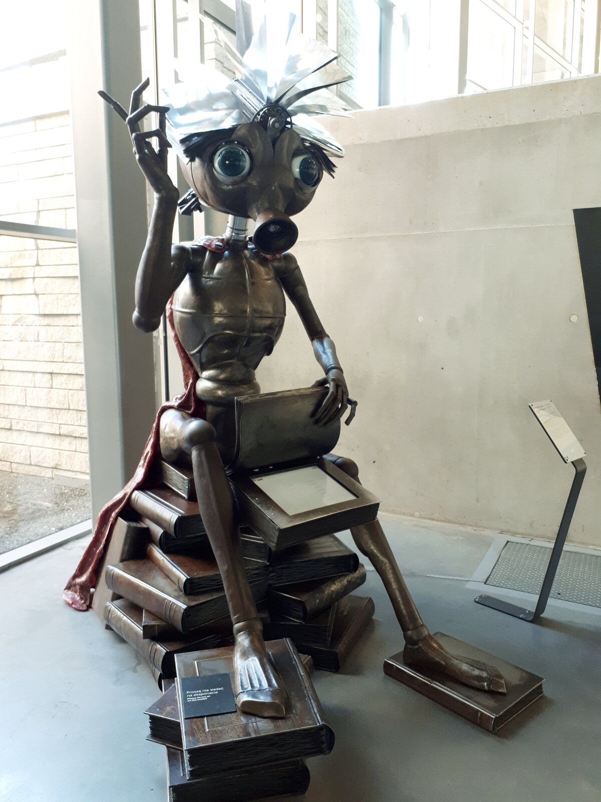 Ilustracja przedstawia rzeźbę postaci - robota. Robot jest wykonany ze złoto - brązowego metalu. Ma duże, wyłupiaste oczy, przypominające ludzkie. Zamiast ust ma zwężenie przypominające rurkę. Na głowie ma otwartą książkę z błyszczącymi, srebrnymi stronami. Jego budowa ciała przypomina budowę człowieka - ma dwie ręce zakończone dłońmi, tułów zwężający się ku dołowi oraz nogi zakończone stopami. Robot ma chude ręce oraz nogi, za kolana i łokcie ma metalowe kule. Na plecach ma czerwoną pelerynę. Robot siedzi na stosie metalowych książek. Jedną z nich ma otwartą, a w środku znajduje się elektroniczny wyświetlacz. W tle znajdują się okna i betonowa ściana.