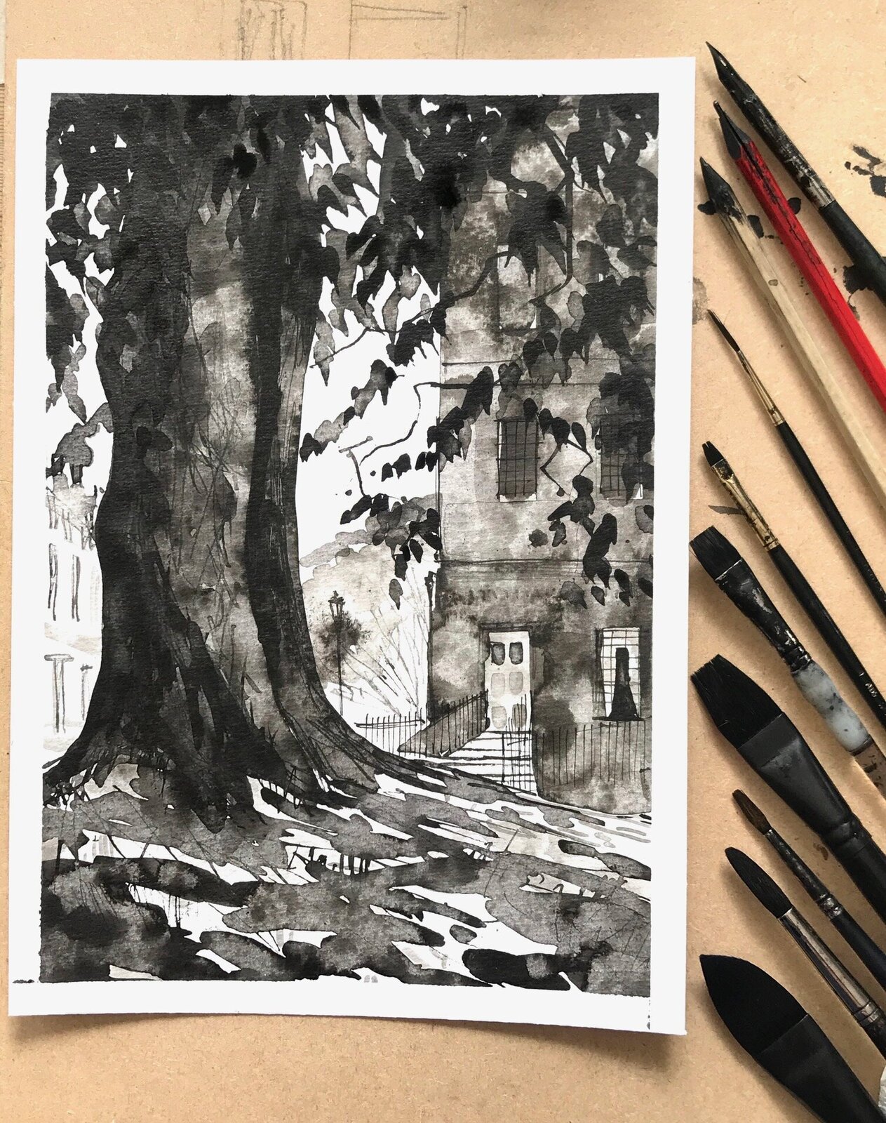 Ilustracja przedstawia rysunek oraz narzędzia, za pomocą których został wykonany - pędzle o różnej grubości i kształcie włosia. Zaprezentowany rysunek przedstawia widok na pień drzewa oraz gałęzie pokryte liśćmi  i budynki mieszkalne. Drzewo znajduje się na pierwszym planie, a za nim, po prawej stronie rysunku stoi jeden z budynków. Drugi budynek stoi po lewej stronie rysunku. Rysunek został wykonany w odcieniach szarości na papierze.