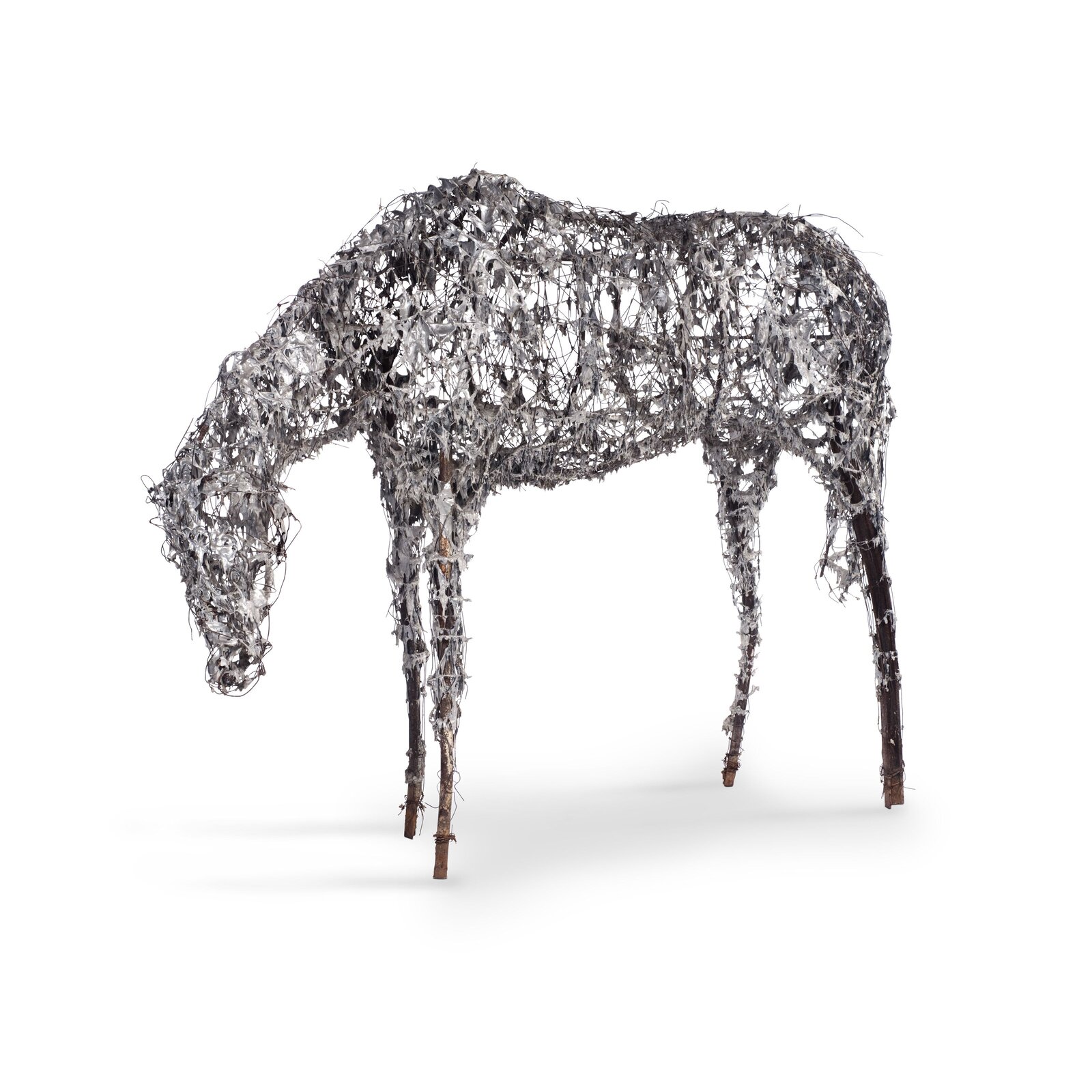 Fotografia przedstawia rzeźbę konia z pochyloną głową, pozbawionego kopyt oraz uszu. Korpus wykonano ze splecionego drutu oraz fragmentów metalu. Kończyny konia wykonano z tyczek oplecionych drutem. Tło, na którym zaprezentowano rzeźbę jest białe.
