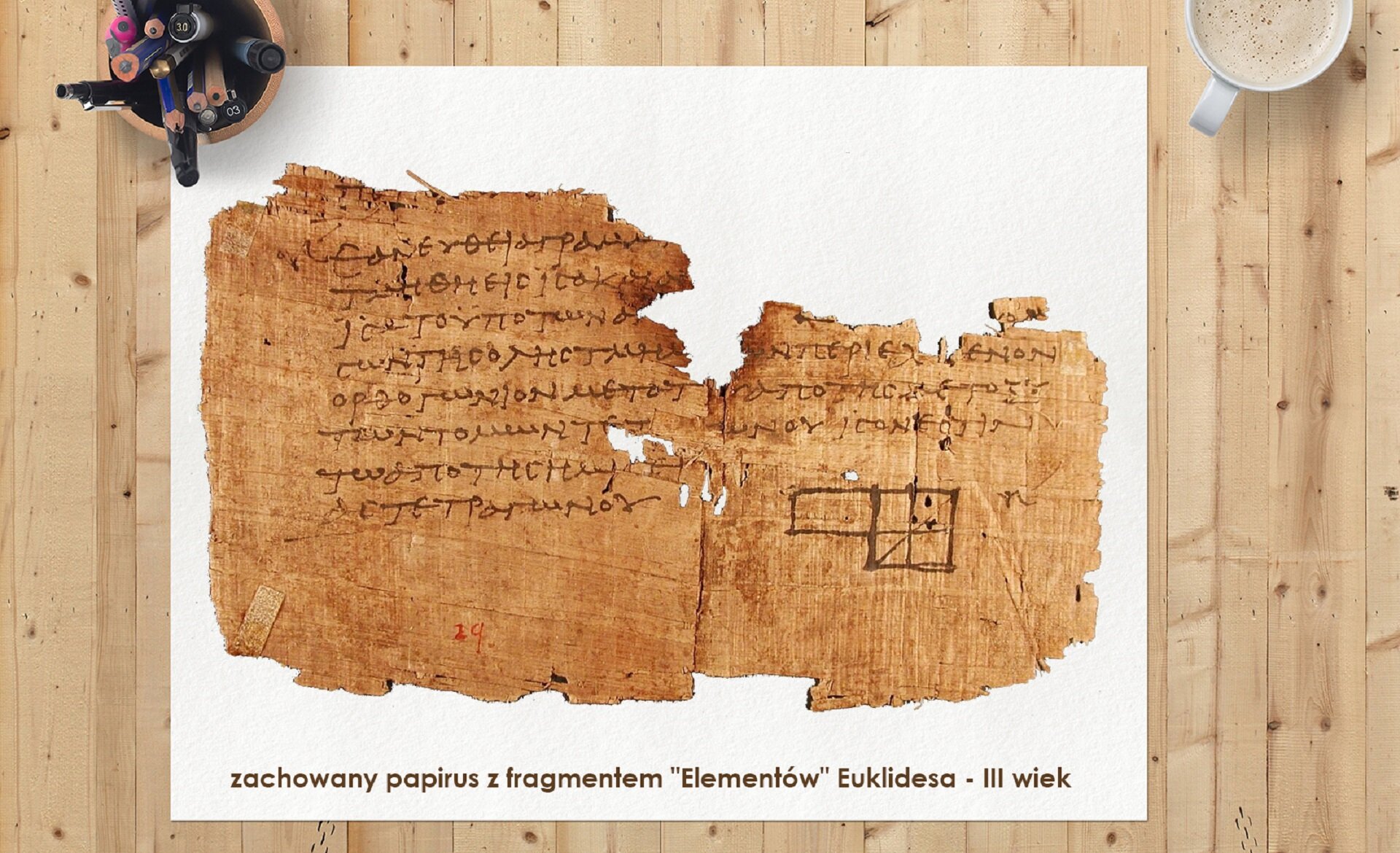 Ilustracja przedstawia sfotografowaną z góry drewnianą płaszczyznę wykonaną z jasnych desek. Na deskach leży biały arkusz. Na nim leży fragment wystrzępionego papirusu. Papirus pokryty jest pismem greckim. U dołu tekstu widać rysunki czworoboków. Na dole białego arkusza widnieje napis informujący czym jest pokazany papirus : „zachowany papirus z fragmentem „Elementów” Euklidesa – III wiek” Lewy górny róg białego arkusza dociśnięty jest do blatu kubkiem, w którym znajdują się kolorowe kredki, długopisy i pisaki. W prawym górnym rogu widać fragment filiżanki z kawą.