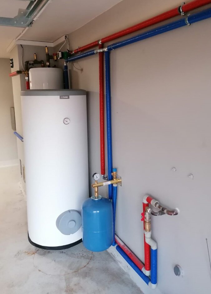 Na zdjęciu przedstawiono wnętrze kotłowni z dużym zbiornikiem na wodę o kształcie walca oraz niewielkim zbiornikiem wyrównawczym. Wzdłuż ściany biegną przewody łączące poszczególne elementy instalacji grzewczej. Przewody te zostały zabezpieczone otuliną w kolorze czerwonym i niebieskim.