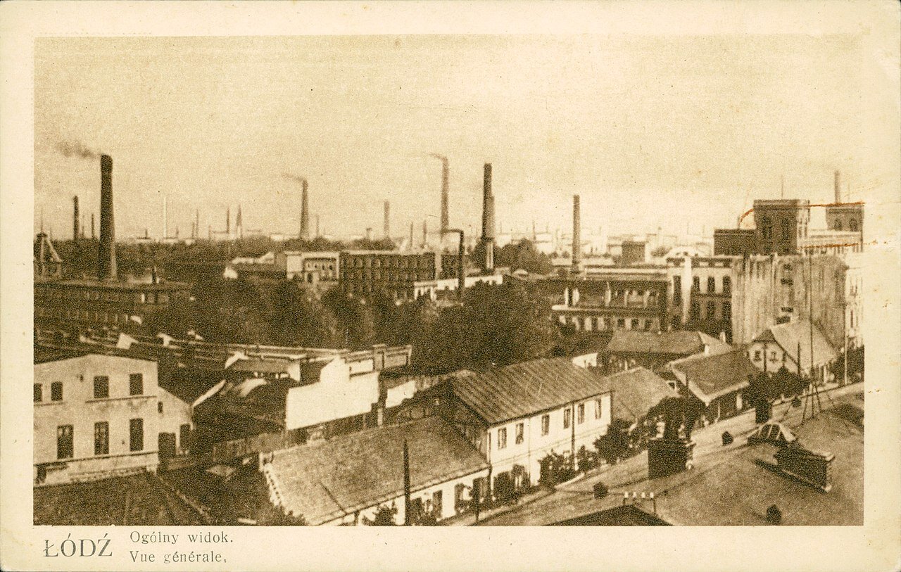 Zdjęcie przedstawia panoramę miasta. Wśród budynków znajdują się wysokie kominy, z których wydobywa się dym.