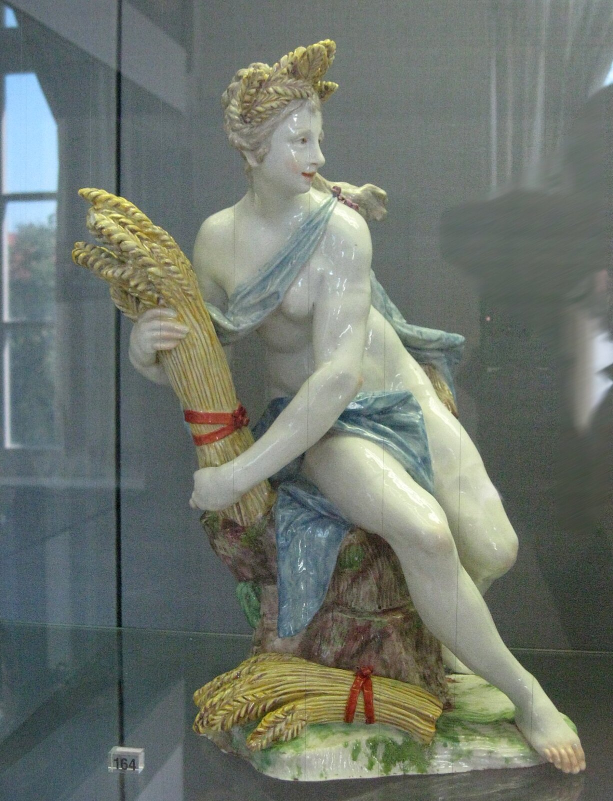 Zdjęcie przedstawia kolorową porcelanową figurkę młodej półnagiej kobiety, która siedzi na pniu. W jasnych włosach wplecione ma snopki zboża, część z nich trzyma również w rękach. Jej skóra jest blada, a nieliczne strzępki szaty, które zakrywają częściowo kobietę są w błękitnym kolorze.