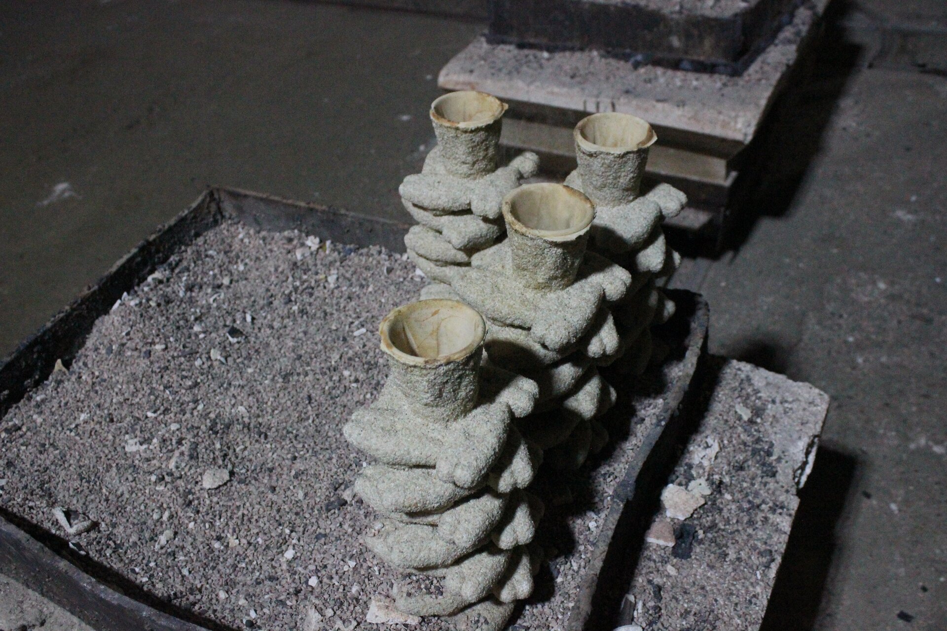 Ilustracja przedstawia cztery formy po wytopieniu z nich woskowych modeli. Formy są podłużne i ustawione otworem wlewowym do góry. Całość umieszczona jest na podstawce z popiołem.