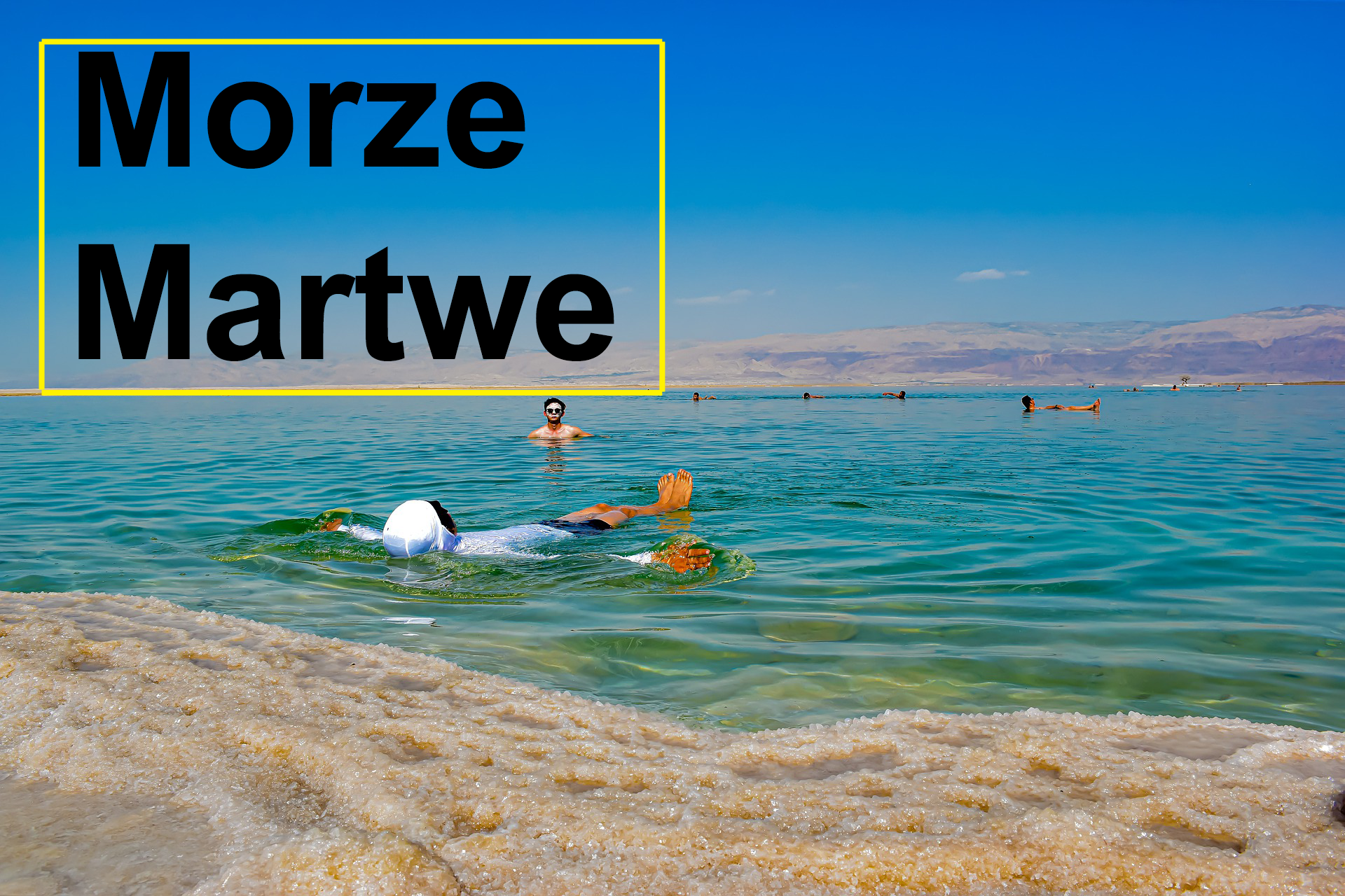 Fotografia przedstawia widok na spokojne morze. Na pierwszym planie piasek wymieszany z morską solą. W centralnym punkcie człowiek w białej bluzie z kapturem na głowie leży, unosząc się na wodzie. W oddali widać więcej sylwetek ludzi zażywających kąpieli. W górnej części błękitne niebo. W lewym górnym roku w żółtej prostokątnej ramce czarny napis: Morze Martwe. 