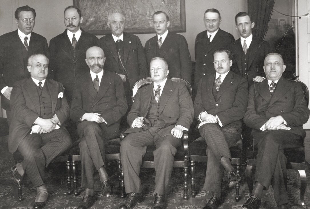 Czarno‑biała fotografia przedstawia członków prezydium BBWR. Mężczyźni ubrani w garnitury pozują do zdjęcia, niektórzy z nich siedzą na krzesłach, a inni stoją za nimi. Za nimi na ścianie wisi obraz. 