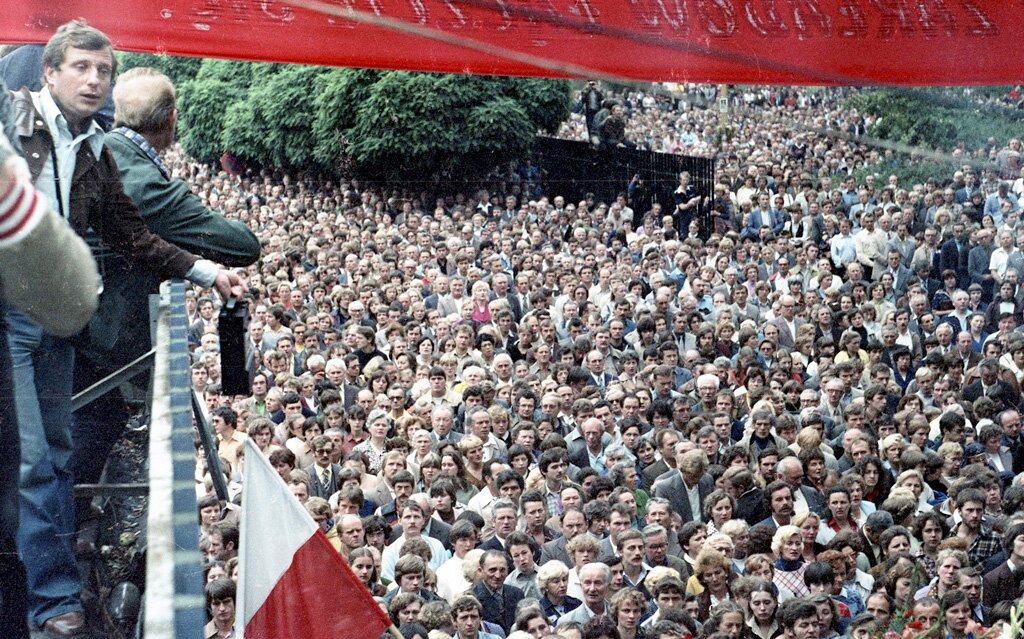 Kliknij, aby powiększyć. Zdjęcie przedstawia setki zgromadzonych ludzi. Po prawej widać ludzi, którzy stoją na podwyższeniu. Powiewa polska biało‑czerwona flaga.