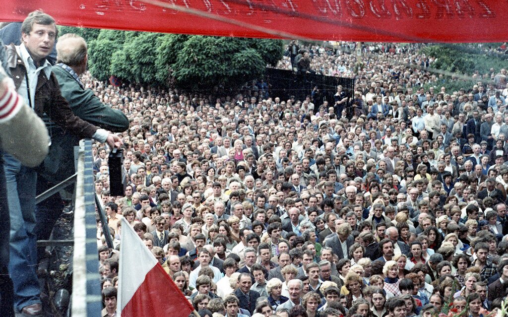 Zdjęcie przedstawia setki zgromadzonych ludzi. Po prawej widać ludzi, którzy stoją na podwyższeniu. Powiewa polska biało‑czerwona flaga. 