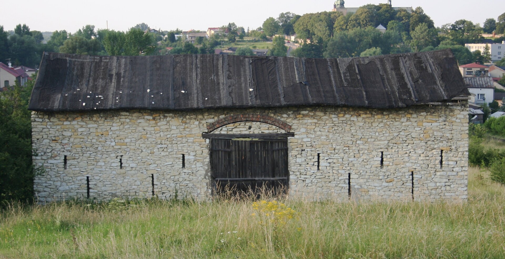 Zdjęcie przedstawia niewielki budynek wykonany z kamienia. Posiada dach wykonany z papy. Brak okien, pośrodku drewniane drzwi. Dookoła teren porośnięty trawą, w tle miasto.