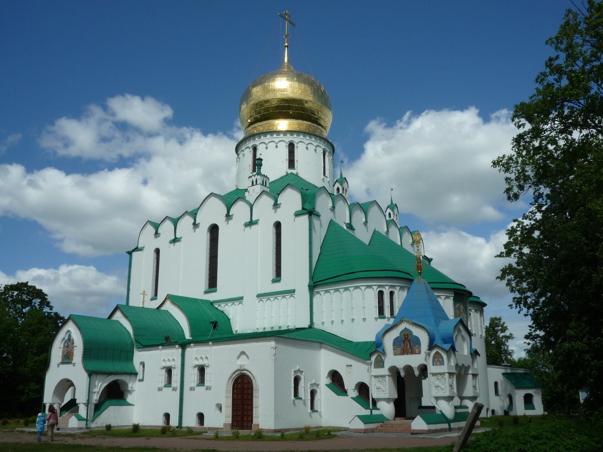 Na zdjęciu cerkiew prawosławna. Biały budynek o rozbudowanej bryle, dach zielony, jeden daszek niebieski przy wejściu od prawej strony, po środku duża złota kopuła na wieży zakończona krzyżem.