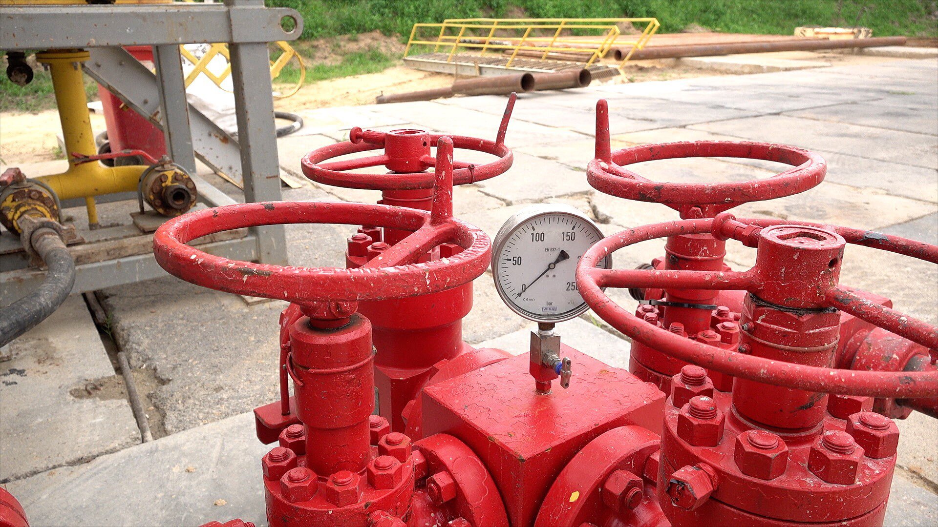 Zdjęcie przedstawia manometr służący do pomiaru ciśnienia. Jest ona zamontowany w części rurociągu. W tle plac wiertni. W górnej części konstrukcji znajduje się okrągła tarcza pomiarowa. Wskazuje ona zero barów. Tarcza osadzona jest w prostopadłościennym, czerwonym korpusie. Posiada on na każdej ze ścian bocznych kołnierze. Dookoła korpusu znajdują się zorientowane pionowo rury, z których każda zakończona jest zaworem wbudowanym w połączenie kołnierzowe. 