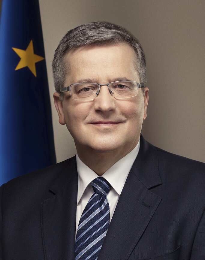 Fotografia przedstawia mężczyznę w średnim wieku w okularach. Ma włosy uczesane na bok i uśmiecha się. W tle widać flagę Unii Europejskiej.