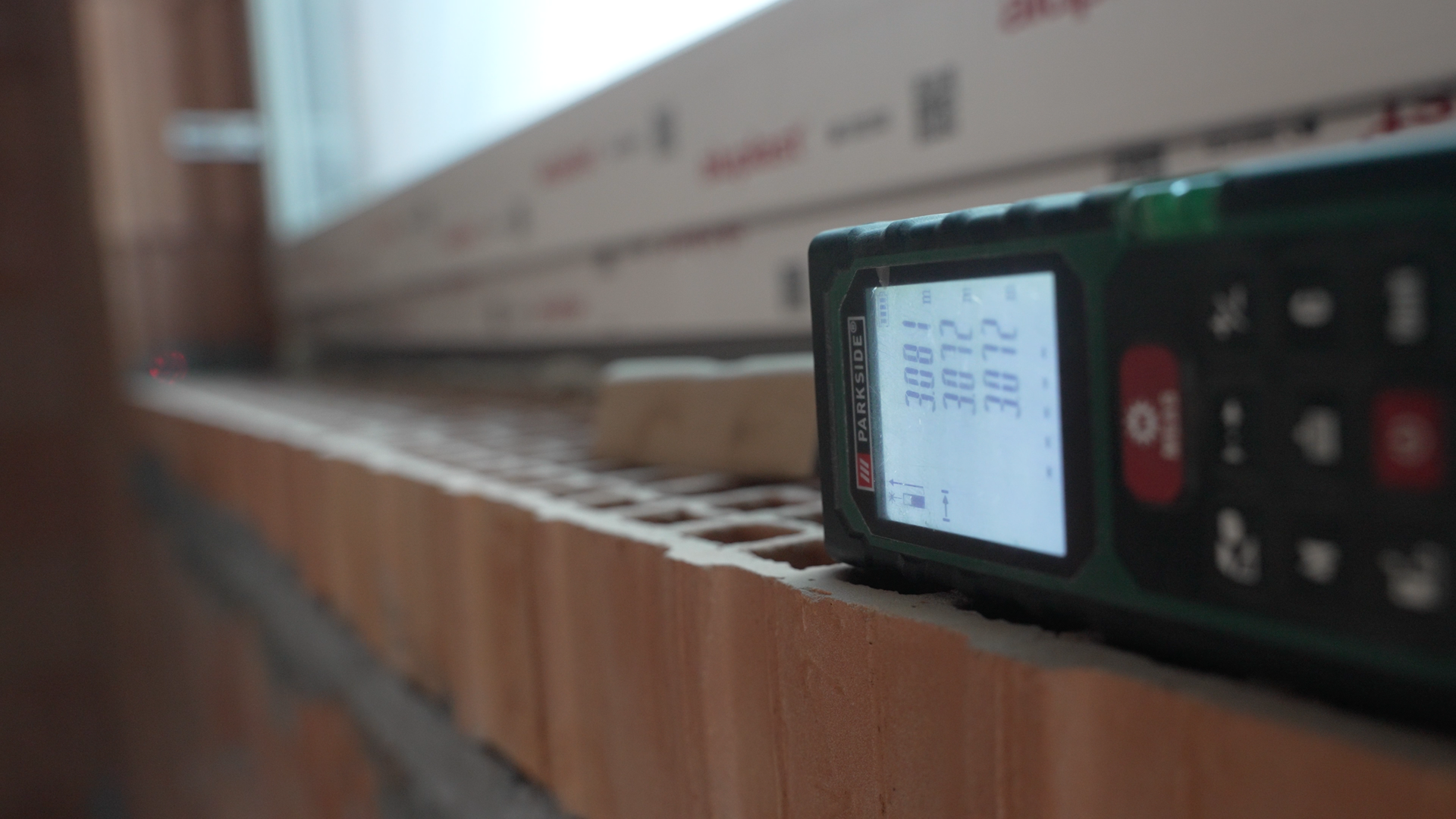 Na ekranie przedstawiono dalmierz leżący na murku obok okna. Na jego ekranie wyświetlają się zmierzone parametry.