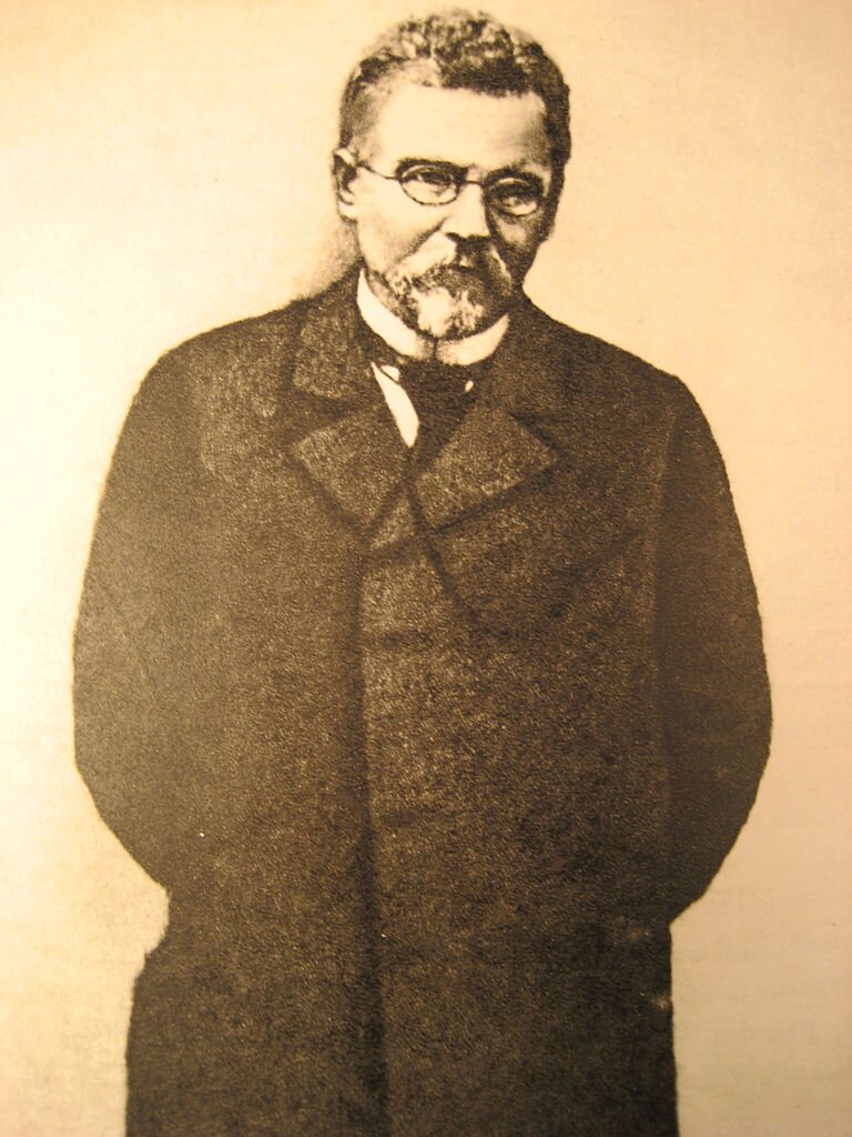 Obraz przedstawia portret dojrzałego mężczyzny. Mężczyzna ma pociągłą twarz. Jego włosy są krótkie, falujące, zaczesane ku górze. Mężczyzna ma niewielką brodę hiszpankę i wąsy. Nosi okulary w drucianej oprawce. Ubrany jest w garnitur, koszulę i krawat. Stoi  z założonymi do tyłu rękami.