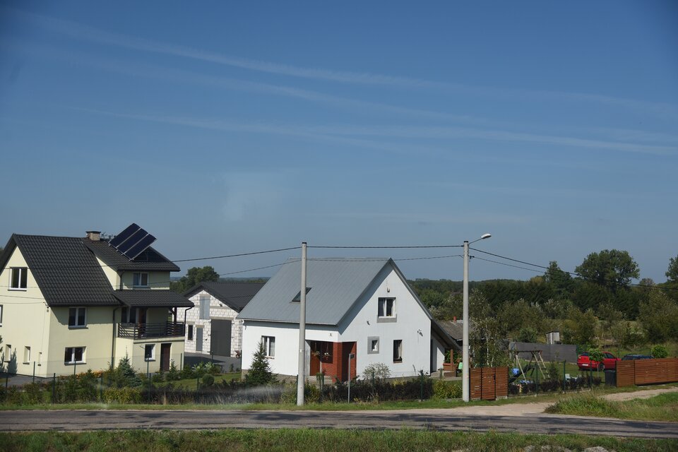 Zdjęcie przedstawia nowoczesne domy. Mają spadziste dachy. Jeden z domów ma panele słoneczne na dachu. Budynki pomalowane są na jasne kolory. Domy są ogrodzone. Stoją przy drodze gruntowej. Za domami widać las.     