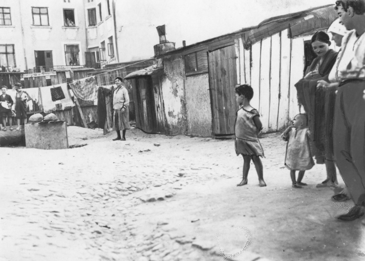 Zdjęcie przedstawiające osiedle bezdomnych ludzi w okresie międzywojennym. Ludzie ci stoją przed prowizorycznymi drewnianymi szopami, przypominającymi dzisiejsze komórki gospodarcze. Na pierwszym planie przed domem stoi rodzina z dwojgiem małych dzieci, na drugim planie stoi mężczyzna. Z tyłu znajduje się duży, ceglany blok.