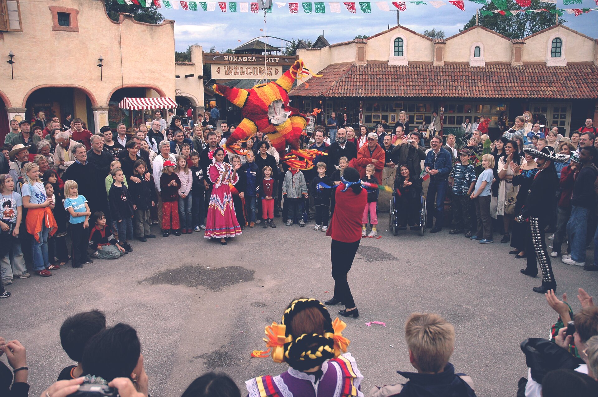  Kolorowe zdjęcie przedstawiające zwyczaj rozbijania piniaty. Na placu grupa ludzi stojąca w kręgu. Na środku zawieszona kolorowa pianiata, pod którą stoją kobieta i mężczyzna w strojach ludowych.