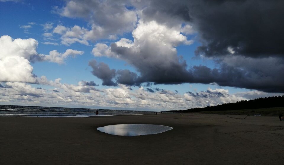 Ilustracja przedstawia fotografię pejzażu morskiego. Większość kadru zajmuje plaża z wielką, okrągłą kałużą pośrodku, w której odbijają się chmury. Błękitne niebo jest pełne chmur od białych aż po czarne - jakby tuz przed burzą.