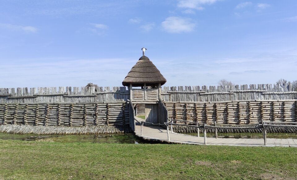 Ilustracja przedstawia rekonstrukcję bramy osady w Biskupinie. W centrum znajduje się drewniana prostokątna brama o masywnej konstrukcji. W dolnej części zbudowana jest z pionowych bali Górną kryje słomiana strzecha. Po obu stronach bramy ciągną się drewniane palisady — wysokie ogrodzenie z zaostrzonych pali, wbitych w ziemię. Dolna ich część składa się z poziomych bali, a górna z pionowych. Do bramy prowadzi drewniany pomost z poręczami. Scena znajduje się na otwartej przestrzeni pod jasnym, błękitnym niebem.  