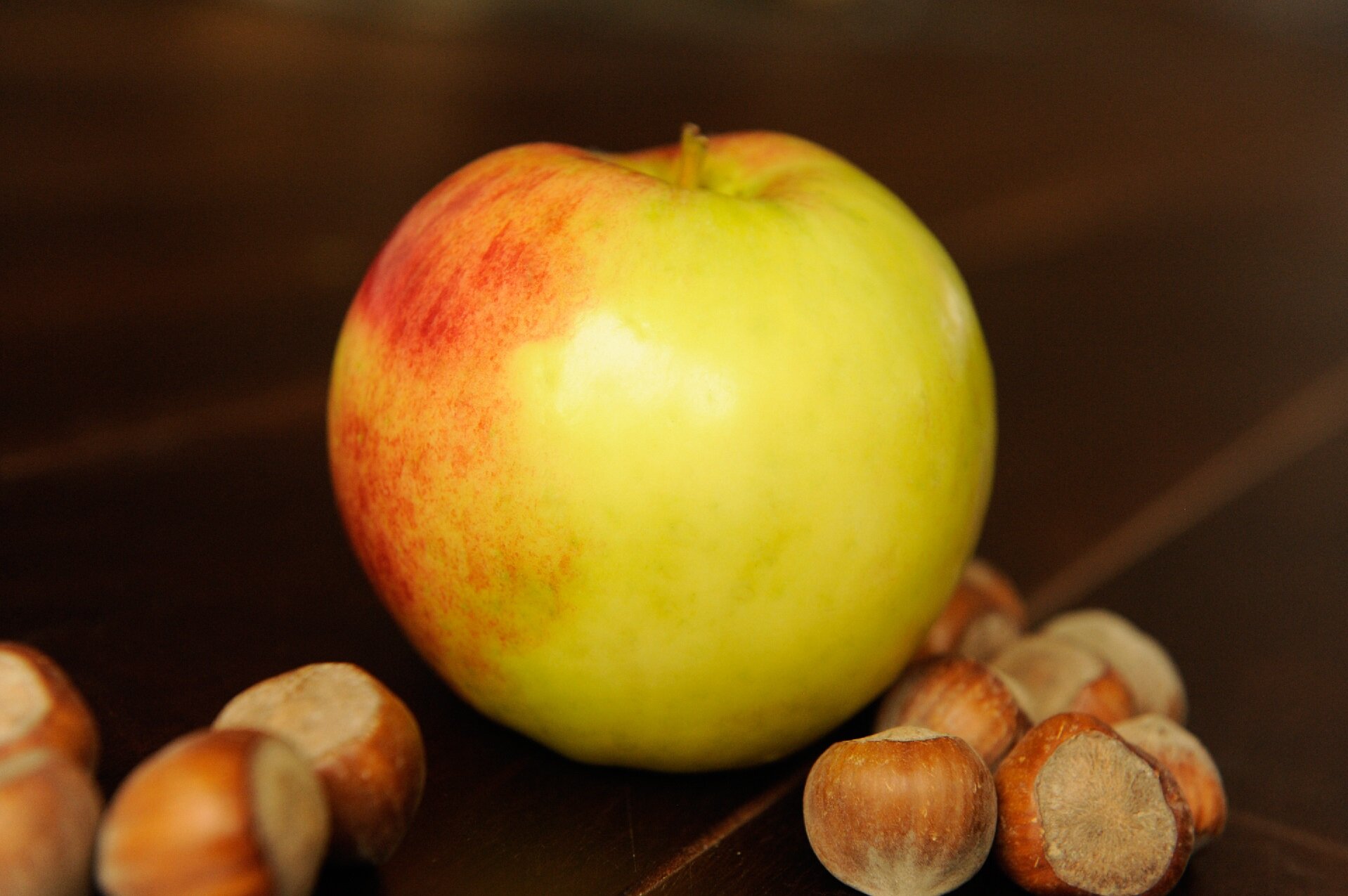 Na fotografii przedstawiono leżące na drewnianym stole dojrzałe jabłko, wokół którego znajdują się rozsypane orzechy laskowe. Skórka na widocznej prawej części jabłka jest żółta, natomiast im bardziej na lewo tym jest bardziej czerwona.  