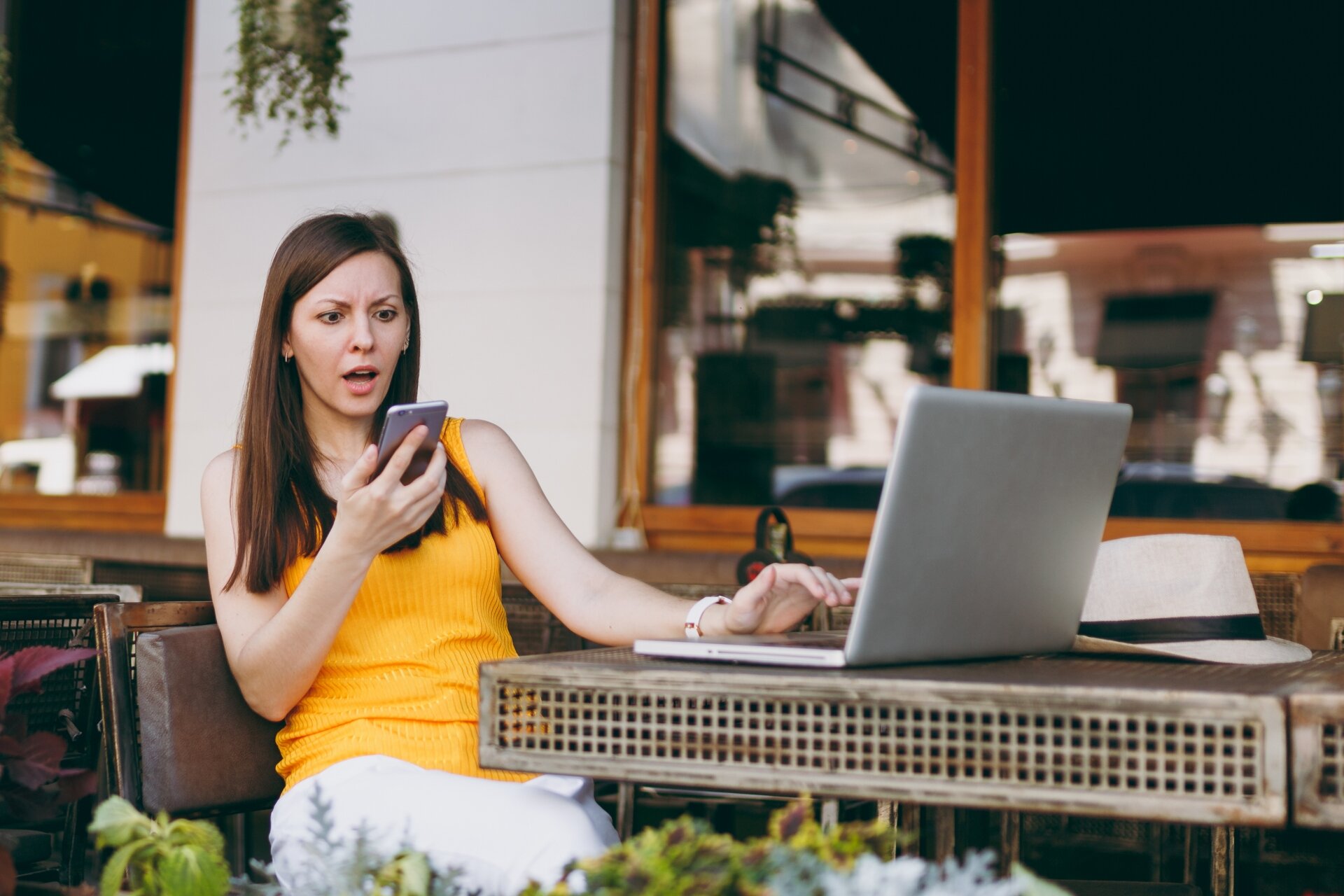 Zdjęcie przedstawia młodą kobietę z długimi rozpuszczonymi włosami, ubraną w jasne spodnie i żółtą bluzkę bez rękawów. Kobieta siedzi w ogródku kawiarnianym. Na stole przed nią stoi laptop. Kobieta w ręku trzyma telefon i ze strachem wpatruje się w jego ekran.
