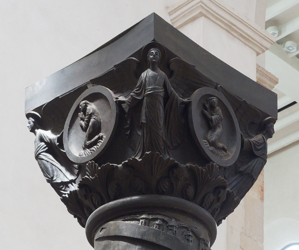 Romański kapitel kolumny z ciemnego metalu lub kamienia, bogato rzeźbiony w postacie ludzkie: centralna stojąca figura w długiej szacie, po bokach mniejsze postacie oraz reliefy w okrągłych polach; całość o masywnej formie, umieszczona we wnętrzu kościoła.