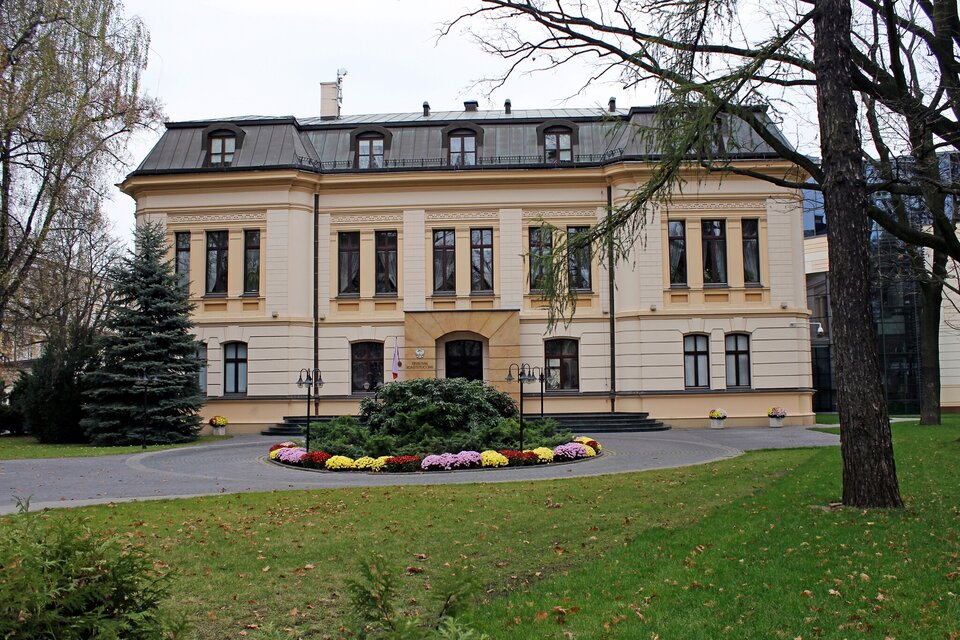 Na zdjęciu znajduje się budynek - siedziba Trybunału Konstytucyjnego. Wyglądem przypomina niewielki pałac z wyeksponowanym wejściem i wysokimi drewnianymi oknami. Budynek jest zmodernizowany i składa się z 2 pięter. Na pierwszym piętrze znajdują się bardzo wysokie okna, zaś na drugim piętrze okna umieszczone są w dachu. Na ostatnim piętrze jest również taras.  Do budynku prowadzi droga, a przed wejściem przyjmuje ona kształt okręgu, na środku którego znajduje się obiekt zielony otoczony ozdobnymi kwiatami.  W tle widoczna jest trawa i liście oraz pojedyncze drzewa iglaste i liściaste.