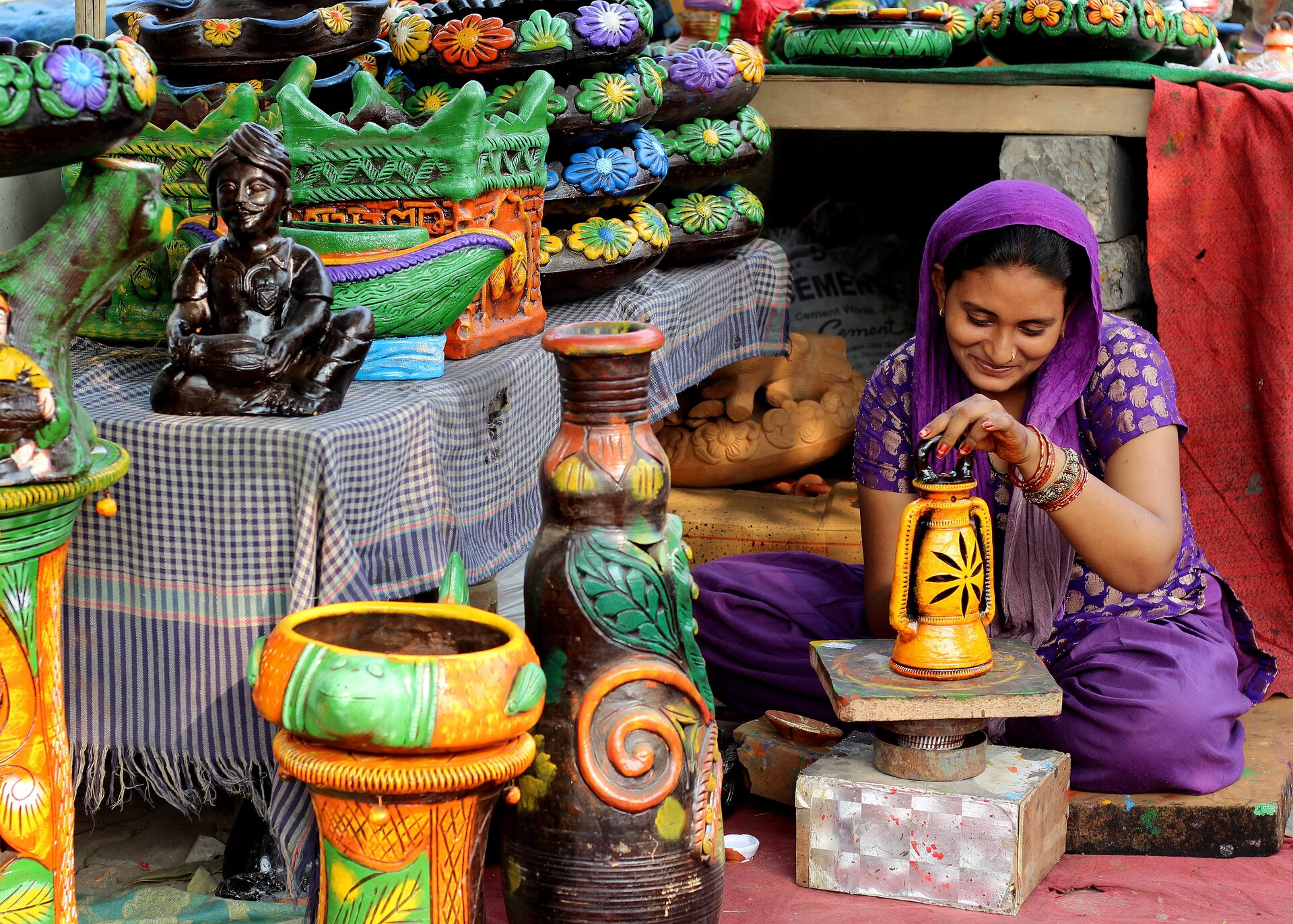 Ilustracja przedstawia hinduskę, która siedzi na ziemi. Kobieta ma czarne włosy, jest uśmiechnięta. Kobieta na lewej ręce ubrane ma bransoletki. Ubrana jest w fioletowy kombinezon a dookoła niej znajdują się ceramiczne elementy m.in. wazony i figurki. Wszystkie elementy są kolorowe. Na zdjęciu dominują kolory pomarańczowy, zielony oraz fioletowy.