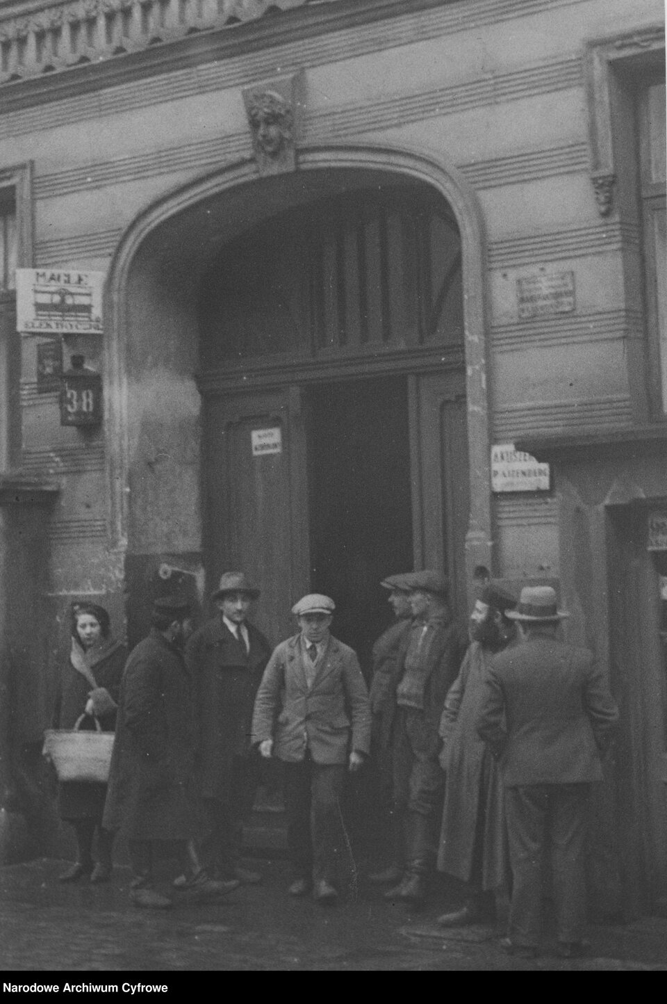 Zdjęcie przedstawia grupę ośmiorga ludzi stojących w bramie kamienicy. Wśród nich znajduje się kobieta w średnim wieku ubrana w płaszcz, trzymająca w lewej ręce koszyk, dwóch Żydów w chałatach, mężczyzna w średnim wieku w płaszczu i kapeluszu, mężczyzna w garniturze i kapeluszu (odwrócony tyłem do widza) i dwóch mężczyzn w marynarkach, spodniach i kaszkietach na głowie. Wysoko, przy bramie znajduje się numer ulicy trzydzieści osiem.