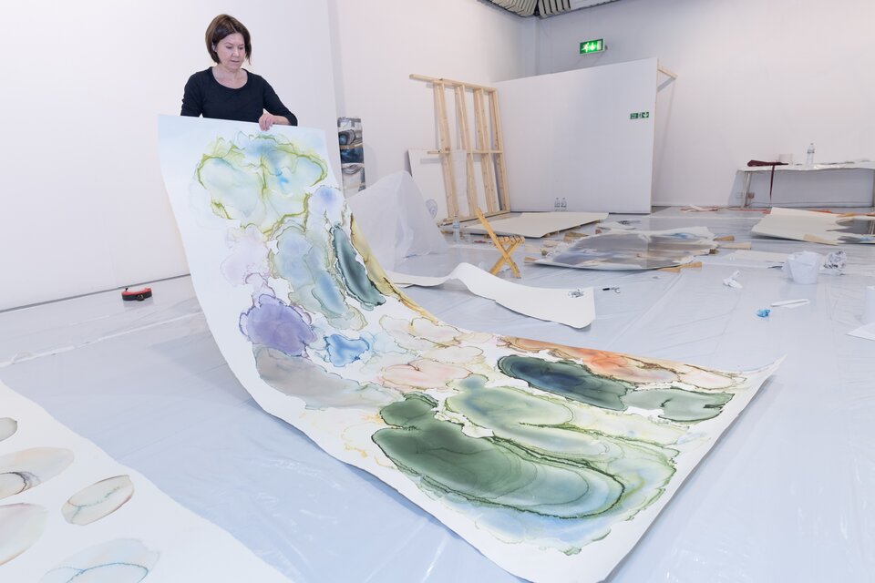 Fotografia przedstawia artystkę oraz jej dzieło. Kobieta ma ciemne włosy sięgające uszu i ubrana jest w czarną bluzkę. Stoi w jasnym pomieszczeniu, w którym na podłodze znajdują się rozłożone prace wykonane techniką akwareli. Artystka rękoma przytrzymuje jedną ze swych prac, to prostokątne, wielkoformatowe dzieło. Praca składa się z dużych, okrągłych i owalnych kolorowych plam, o rozmaitym stopniu intensywności, kształtem przypominających chmury. Plamy są głównie w odcieniach zieleni, niebieskiego i fioletu, mają miękkie krawędzie i w większości widoczne kontury. Niektóre kształty są intensywniejsze i zawierają więcej barwnika, a niektóre są blade, rozbielone. 