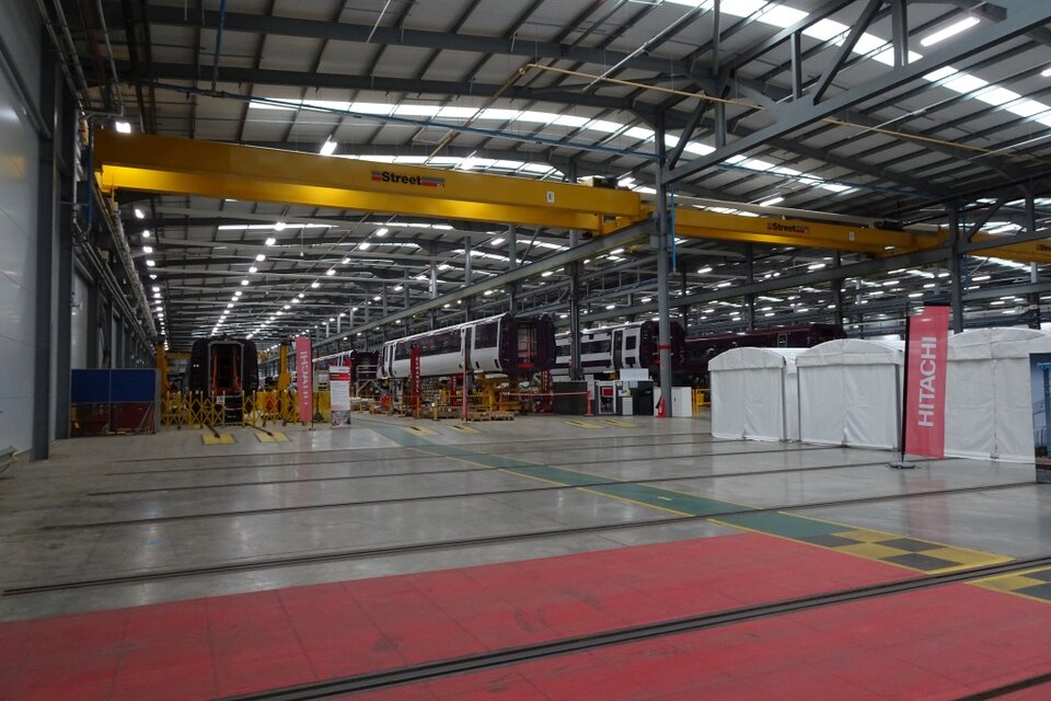  Wnętrze nowoczesnej hali produkcyjnej taboru kolejowego. Po obu stronach szerokiego przejścia stoją na żółtych podporach dwa białe składy pociągów w trakcie montażu, ustawione symetrycznie przodami do obserwatora. W centrum kadru idzie pracownik w ciemnym ubraniu. Hala jest wysoka, jasna, równomiernie oświetlona, z widoczną konstrukcją dachu i torami w posadzce, co podkreśla przemysłowy charakter zakładu.