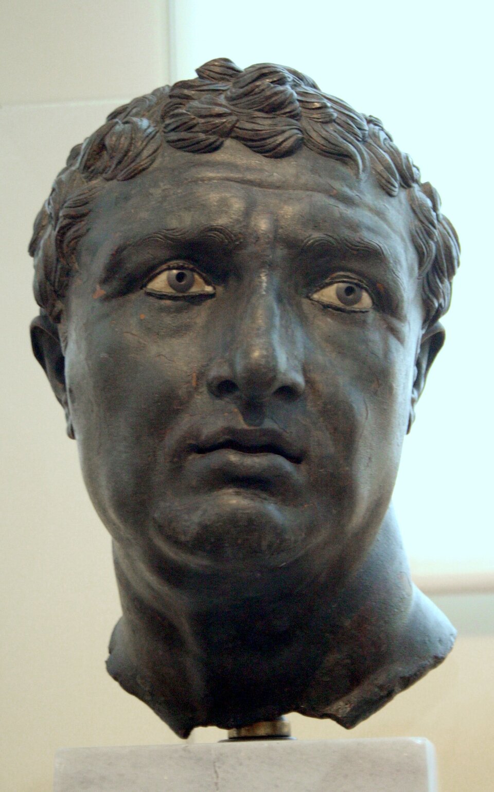 Ilustracja przedstawia brązowy portret nieznanej osoby z I wieku p.n.e., przechowywany w Narodowym Muzeum Archeologicznym w Atenach. Widzimy realistycznie oddaną głowę mężczyzny o krótkich, falujących włosach i wyrazistych rysach twarzy. Oczy mają inkrustowane źrenice, co nadaje spojrzeniu intensywność i głębię. Usta są lekko rozchylone, a policzki i broda modelowane z dużą dbałością o szczegóły anatomiczne. Powierzchnia brązu jest gładka, z delikatnymi przebarwieniami wynikającymi z wieku. Głowa ustawiona jest na prostym kamiennym cokole, a jednolite tło podkreśla formę rzeźby.