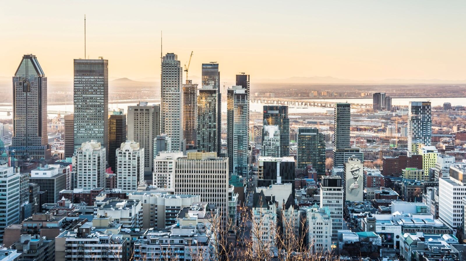 Ilustracja przedstawia panoramę współczesnego miasta. Zabudowa jest gęsta i składa się z wieżowców o różnej wysokości i kształcie. W oddali widać rzekę oraz jasne niebo rozświetlone blaskiem wschodzącego słońca. 